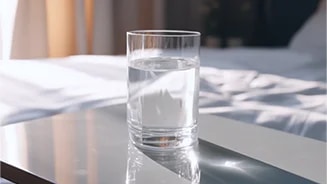 투명한 유리컵에 물이 담겨 있으며, 컵은 매끄러운 표면 위에 놓여 있습니다. 배경에는 부드러운 천이 드리워져 있고, 자연광이 들어와 밝은 분위기를 연출하고 있습니다.