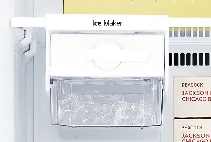 냉장고 내부의 아이스 메이커가 보입니다. 아이스 메이커 위에는 Ice Maker라는 텍스트가 적혀 있습니다. 아이스 메이커 아래에는 투명한 용기에 얼음이 담겨 있습니다. PEACOCK JACKSON CHICAGO B라는 텍스트가 적힌 상자가 보입니다.
